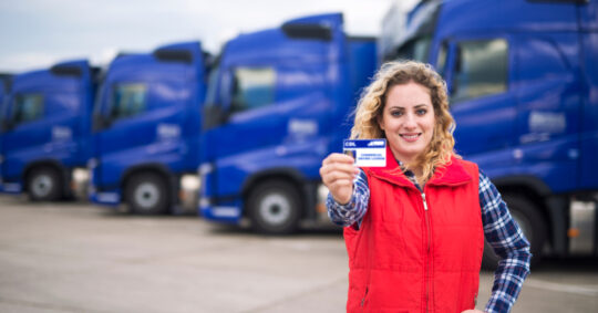 woman holding cdl license in parking lot with semi trucks